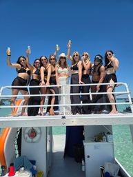 Group of women in bikinis and cover-ups raising canned drinks on the upper deck of a boat, celebrating over clear turquoise water beneath a bright blue sky.