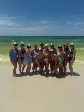 Group of women in colorful swimsuits and sun hats posing at the water’s edge on a white-sand beach with turquoise ocean and a clear blue sky.