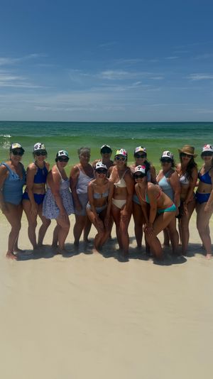 Group of women in colorful swimsuits and sun hats posing at the water’s edge on a white-sand beach with turquoise ocean and a clear blue sky.