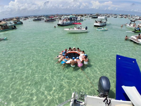 Crowded tropical sandbar with dozens of anchored boats and pontoons, people partying on a round inflatable ring in crystal-clear shallow turquoise water