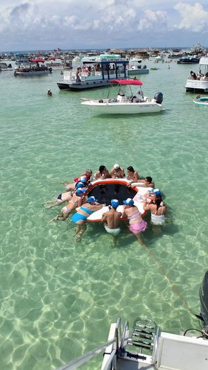 Crowded tropical sandbar with dozens of anchored boats and pontoons, people partying on a round inflatable ring in crystal-clear shallow turquoise water