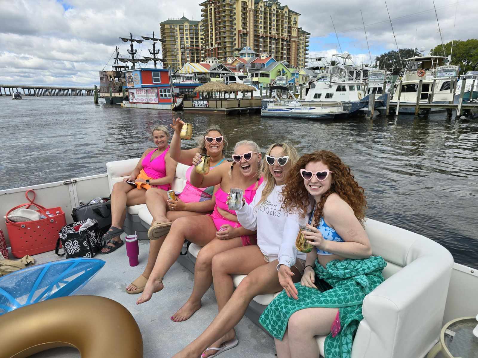 Boat party with five women in bright swimsuits and heart-shaped sunglasses toasting drinks on a motorboat in a busy marina with colorful waterfront buildings, docked yachts, and a tall resort skyline