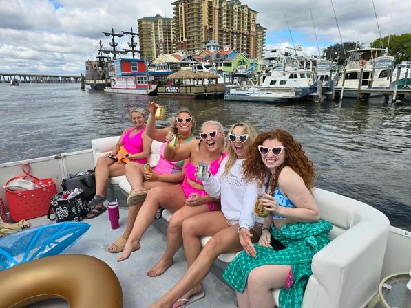 Boat party with five women in bright swimsuits and heart-shaped sunglasses toasting drinks on a motorboat in a busy marina with colorful waterfront buildings, docked yachts, and a tall resort skyline