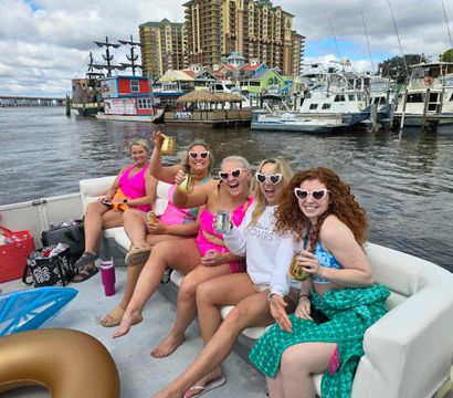 Boat party with five women in bright swimsuits and heart-shaped sunglasses toasting drinks on a motorboat in a busy marina with colorful waterfront buildings, docked yachts, and a tall resort skyline