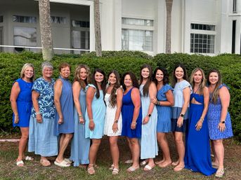 Thirteen women in coordinated blue and white summer dresses smiling and posing side-by-side in front of green hedges, palm trees, and a white coastal-style building — group photo at a tropical courtyard.
