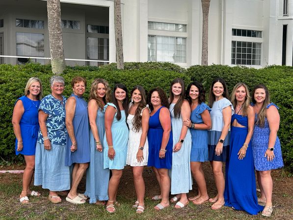 12 women of varying ages wearing coordinated blue dresses and one in white, smiling and posing side-by-side outdoors in front of a hedge, palm trees, and a white coastal-style building.