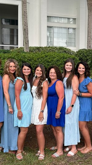 12 women of varying ages wearing coordinated blue dresses and one in white, smiling and posing side-by-side outdoors in front of a hedge, palm trees, and a white coastal-style building.