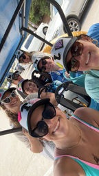 Seven smiling women in sunglasses and decorated caps take a cheerful selfie while riding a blue-roofed golf cart in a sunny parking lot.