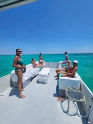 Smiling group of adults relaxing on a pontoon boat in clear turquoise water under a bright blue sky, sunbathing with sunglasses and drinks — carefree day on the ocean.