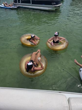 Three people in black swimsuits and caps lounging on gold inflatable tubes, sipping canned drinks while floating in green lake water beside a pontoon boat — summer boating scene.