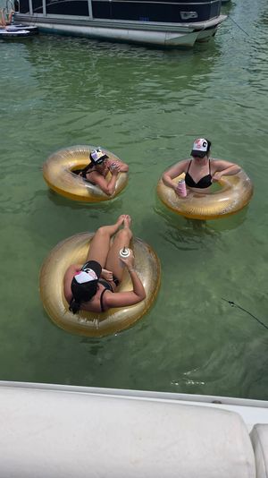 Three people in black swimsuits and caps lounging on gold inflatable tubes, sipping canned drinks while floating in green lake water beside a pontoon boat — summer boating scene.