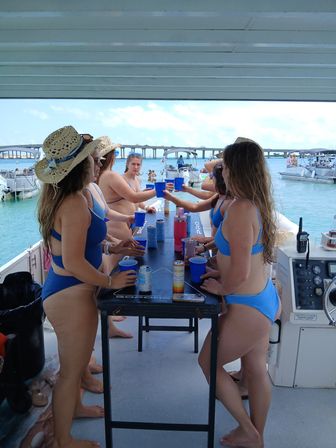 Group of women in blue bikinis toasting with plastic cups around a table on a pontoon party boat over turquoise water with a low bridge and other boats in the background.