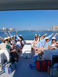 Smiling women in colorful caps and casual beachwear on a sunny boat party, holding drinks and waving against a calm blue bay with waterfront condos and a marina on the horizon