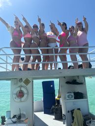 Eight women in pink swimsuits, one wearing a 'Bride' top, cheering and pointing from the upper deck of a boat over clear turquoise water on a sunny bachelorette boat party.