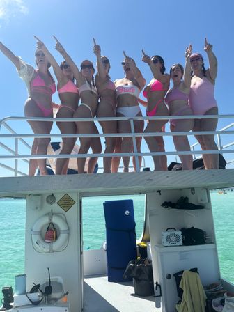 Group of women in pink swimsuits, including a bride, cheering and pointing from a boat deck over turquoise water under a clear blue sky