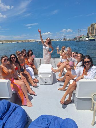 Sunny summer boat party — group of friends on a pontoon boat holding drinks around a cooler on deck, blue sky above with sandy shoreline and marina with sailboats in the background