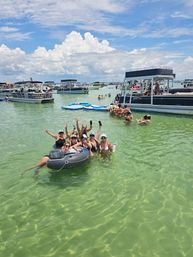 Friends partying on an inflatable tube and wading at a shallow sandbar with anchored pontoon boats, floating mats, and clear green water under a sunny blue sky.