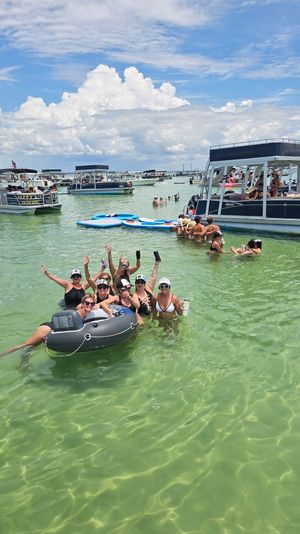 Group of people enjoying a lively sandbar boat party in clear shallow green water, lounging on an inflatable tube and nearby pontoons with anchored boats under a bright blue sky and puffy clouds.