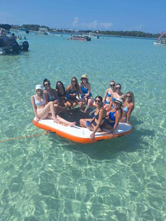 Group of friends in swimsuits lounging on an orange-and-white inflatable platform in crystal-clear shallow turquoise water with boats and a distant sandy shoreline on a sunny beach day.