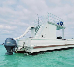 White two-level pontoon boat with rooftop slide and upper deck floating in clear turquoise water, rear outboard engine and swim ladder visible