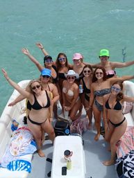 Smiling group of women in bikinis posing on a pontoon boat over clear turquoise water, wearing sunglasses and colorful hats with arms raised for a sunny tropical boat party.