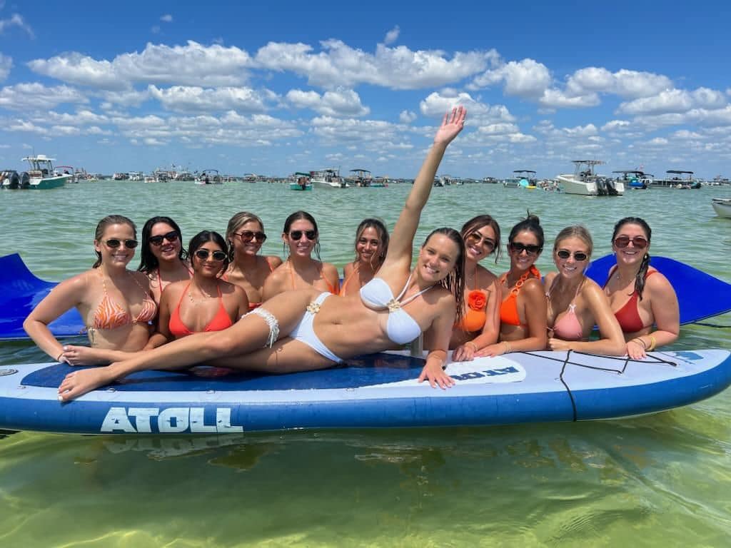 Group of women in colorful bikinis lounging on a large paddleboard at a sunny sandbar in shallow turquoise water with anchored boats and a blue sky with fluffy clouds