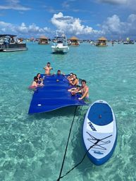 Group relaxing on a large blue floating mat beside a paddleboard in crystal-clear turquoise water, surrounded by boats and tiki-style floating huts at a sunny tropical sandbar.