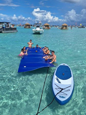 Friends enjoying a sunny sandbar party on a large blue floating mat and paddleboard in crystal-clear turquoise water, surrounded by boats and thatched floating huts.