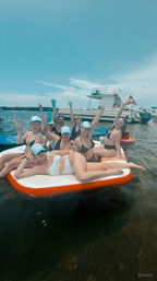 Group of friends in swimsuits and matching light-blue caps lounging and cheering on an orange-and-white inflatable platform in shallow coastal water, holding drinks with a white party boat and American flag in the background under a bright blue sky.