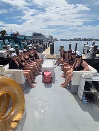 Group of friends in swimsuits seated along benches on a pontoon boat at a marina, cooler and pink bag center, golden inflatable tube visible, docks and coastal skyline under a sunny, partly cloudy sky