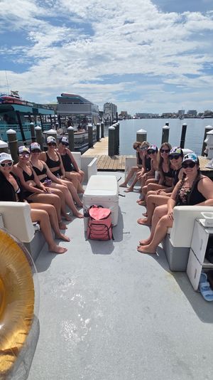 Smiling friends in swimsuits and caps seated on a pontoon-style boat for a sunny marina boat party, docks and waterfront skyline beyond.