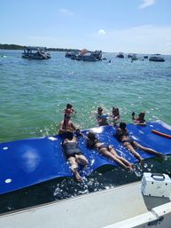 Sunny boat party with friends lounging on a large blue floating mat beside a dock in clear green water, pontoons and party boats clustered in the distance under a bright blue sky