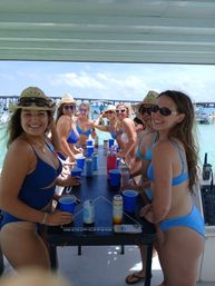 Boat party with friends in blue bikinis and straw hats playing beer-pong on a pontoon-style table over clear turquoise water, anchored boats and a bridge visible in the sunny background