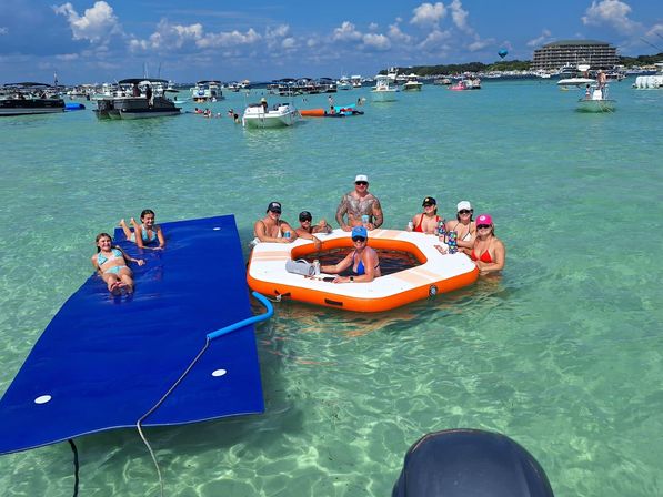 Friends relaxing on an orange inflatable swim platform and blue floating mat in clear turquoise shallow water, surrounded by anchored boats under a sunny sky