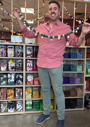 Smiling person holding a colorful felt-ball garland in a craft store, standing before cubbies filled with organized colorful craft supplies.