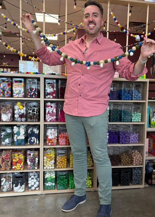 Smiling person holding a colorful felt-ball garland in a craft store, standing before cubbies filled with organized colorful craft supplies.