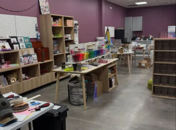 Craft studio interior with purple walls, wooden shelving and work tables displaying colorful art and craft supplies, pens, paper, storage bins and boxes on a concrete floor.