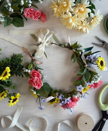 DIY spring floral wreath flatlay with pink roses, yellow sunflowers, small blue and white blossoms, greenery, ribbons and craft scissors on a neutral linen backdrop