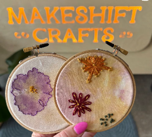 Hand holding two small wooden embroidery hoops featuring pastel-dyed fabric with hand-stitched floral designs in purple, gold, and burgundy, against a blurred bright orange craft sign in a home studio setting.