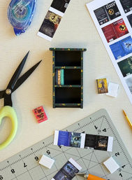 Miniature painted bookshelf and tiny book covers on a cutting mat with scissors, adhesive tape, craft knife and printed book jacket sheets — DIY miniature book craft workspace