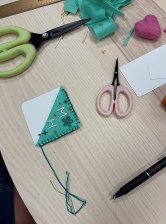 DIY teal felt corner bookmark with embroidered initials and tassel on a light wood craft table surrounded by pink and green scissors, ruler, felt scraps and a black pen