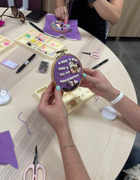 Hands working on a DIY bead-embroidery hoop with purple fabric, arranging pearls, letter beads and floral embellishments at an indoor craft workshop table with trays of beads, scissors and pens.