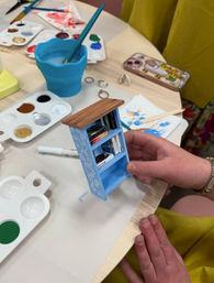 Hand holding a painted blue miniature bookshelf filled with tiny books on a craft table with paint palettes, brushes, a water cup and phone — DIY miniature bookshelf painting project