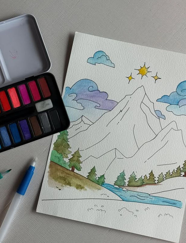 Top-down view of a watercolor mountain landscape on textured paper featuring pine trees, a blue stream, purple clouds and a sun, with an open watercolor palette and water brush pen beside it.