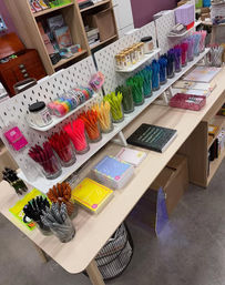 Colorful stationery store display with rainbow pens in glass jars, rolls of washi tape, sticky notes and notebooks neatly arranged on pegboard shelves.