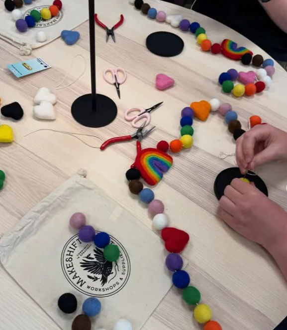 DIY craft workshop table with hands stringing colorful felt balls and rainbow heart shapes into garlands, pliers, scissors, and fabric pouches on a light wood surface.