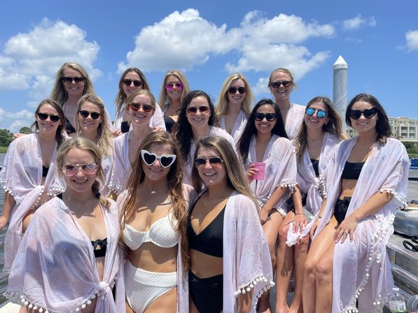Group of women in swimsuits and matching light pom-pom cover-ups and sunglasses posing and smiling on a sunny boat at a marina under a blue sky with fluffy clouds