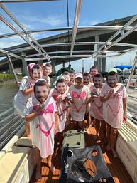 Boat party at a marina under a concrete bridge, people in matching pink bikini-print cover-ups holding identical male face cutouts on a sunny dockside outing