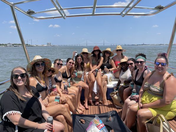 About 15 people in swimsuits and sun hats enjoying drinks on a sunny boat cruise, seated on a wooden deck with a coastal skyline and suspension bridge in the background.