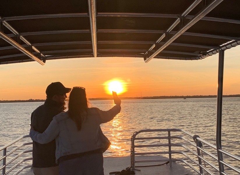 Silhouetted couple embracing on a covered boat deck taking a sunset selfie as the golden sun reflects across calm water